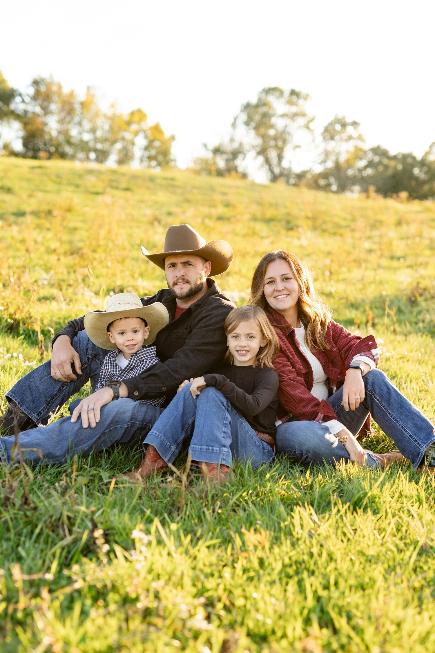 A happy family of four wearing cowboy hats sits together on a sunny green grassy hillside at golden hour.