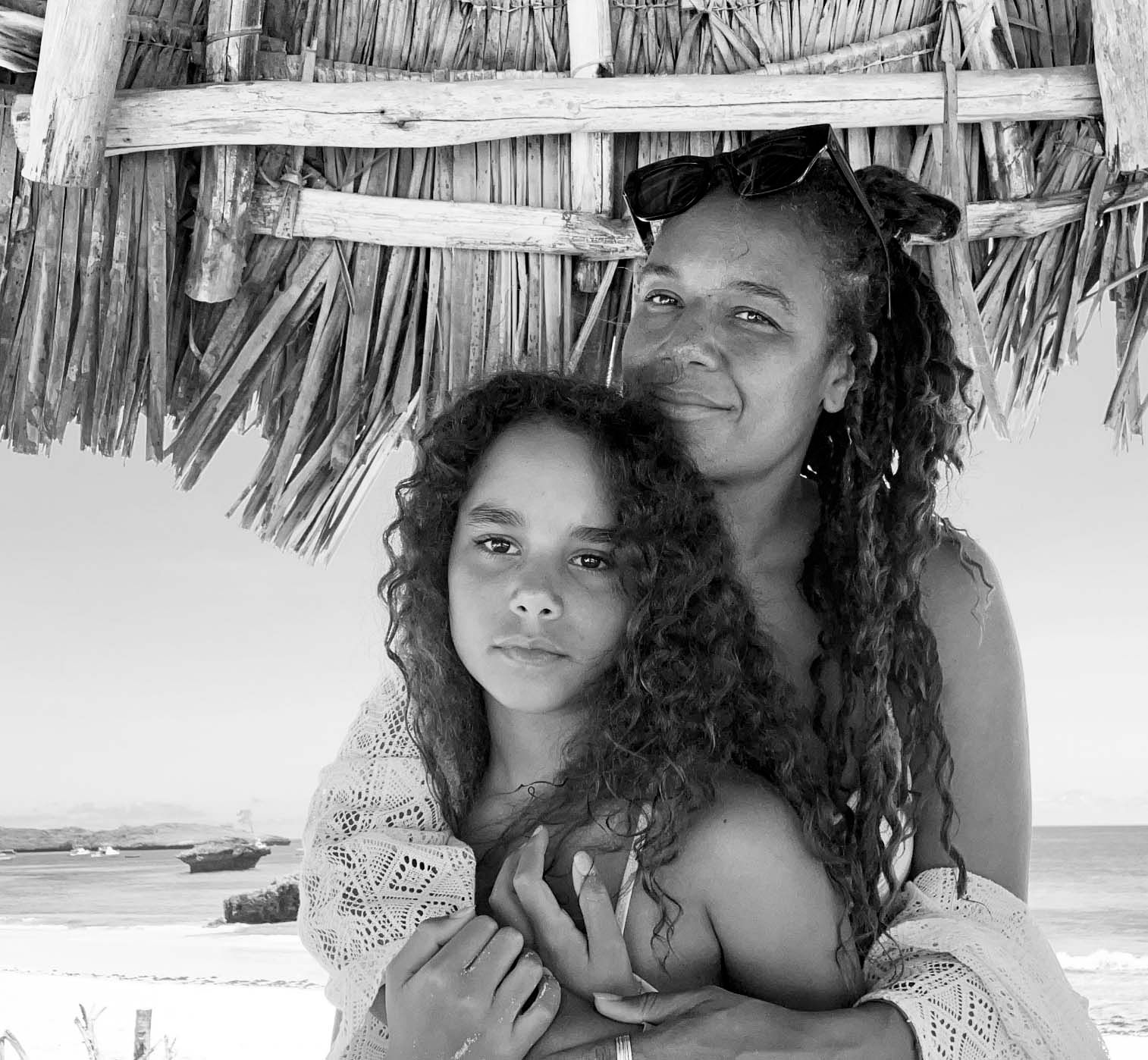 A black and white portrait of a parent and child with curly hair embracing under a thatched beach shelter.