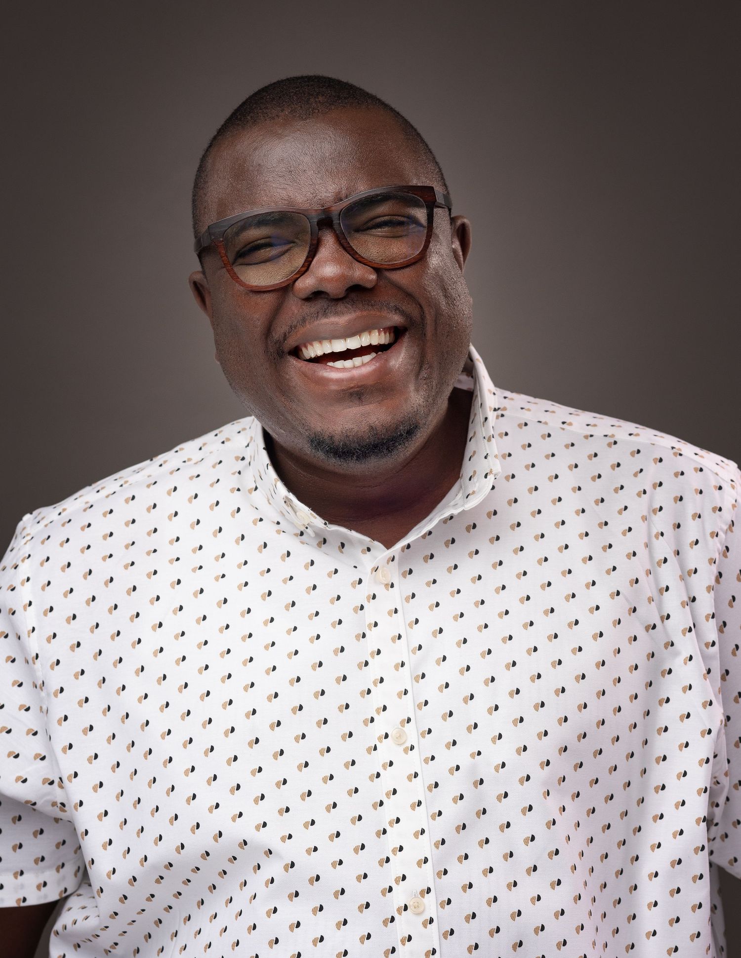 A person wearing glasses and a white patterned shirt smiles warmly against a dark background.
