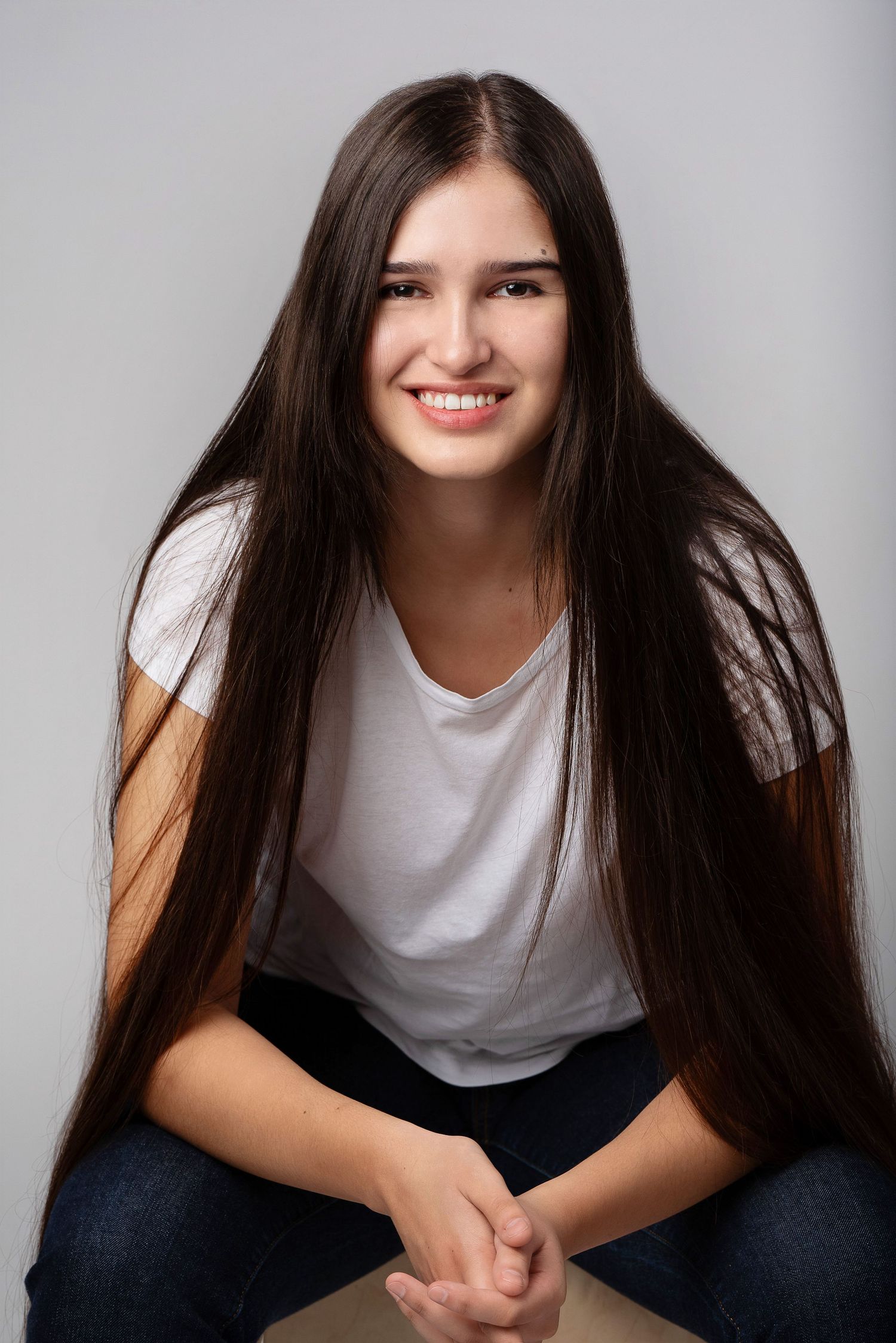 Person with long dark hair wearing a white t-shirt and jeans sits casually against grey background in studio portrait.