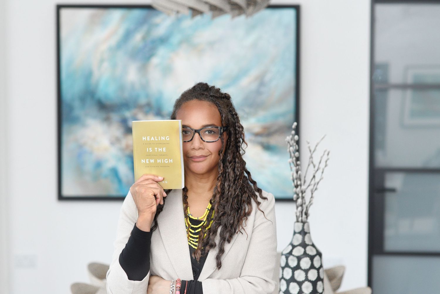 Someone wearing glasses and a white blazer holds up a yellow book while sitting in an office setting.