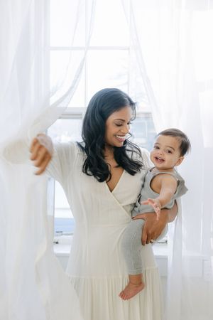 Smiling mother in white dress holding happy baby by bright window with sheer curtains.