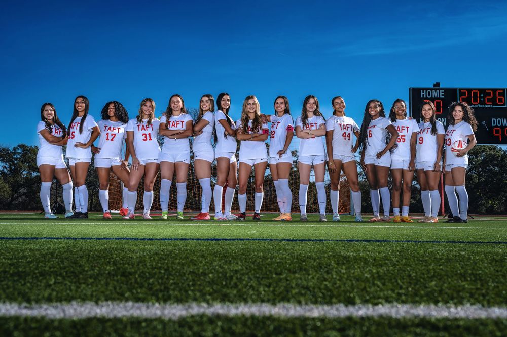 Group photo of softball team members wearing white uniforms lined up on a green field with blue sky background.