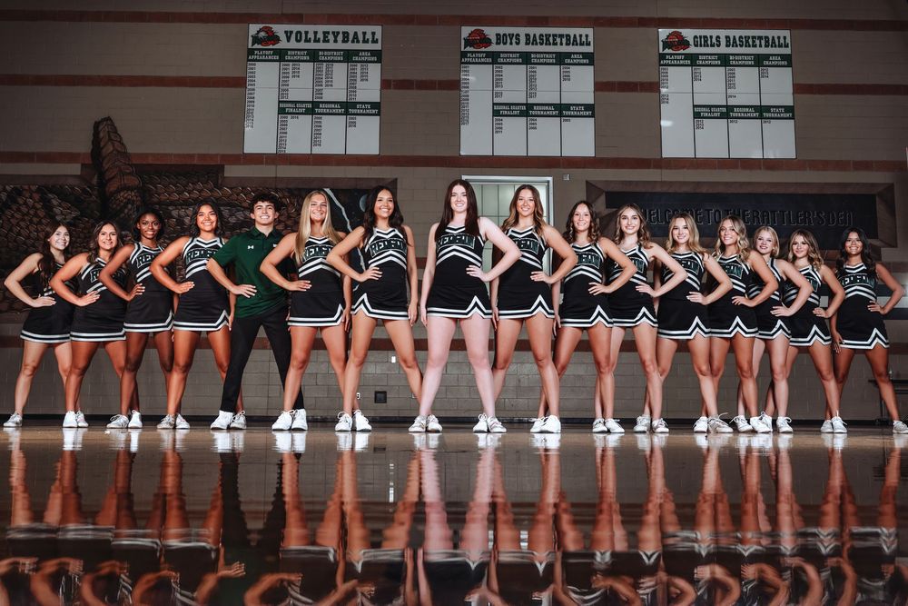 A team poses together in uniform black and white outfits on a reflective wooden basketball court surface.