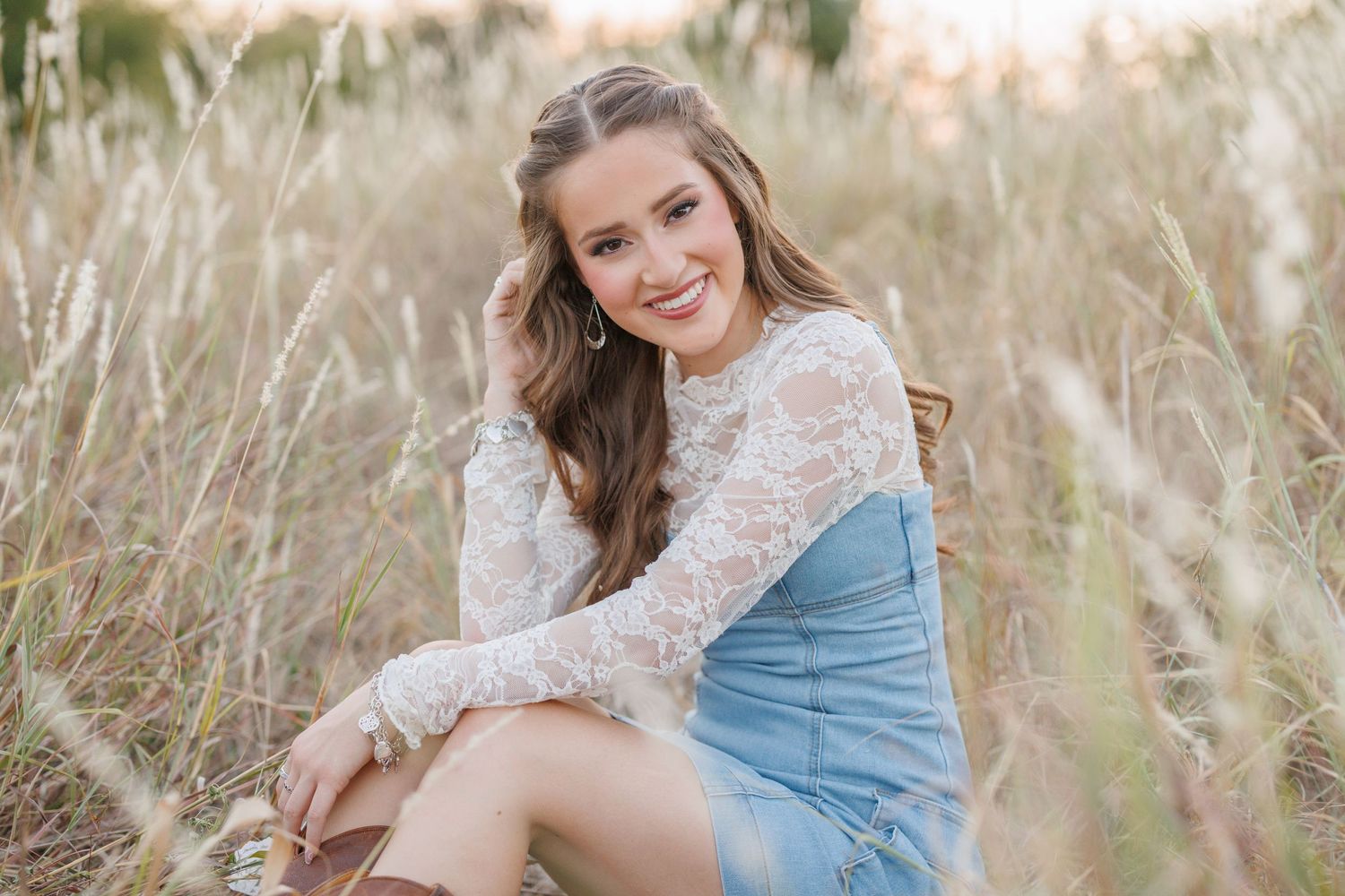 Outdoor portrait in field wearing white lace top and blue dress among tall grasses at sunset.