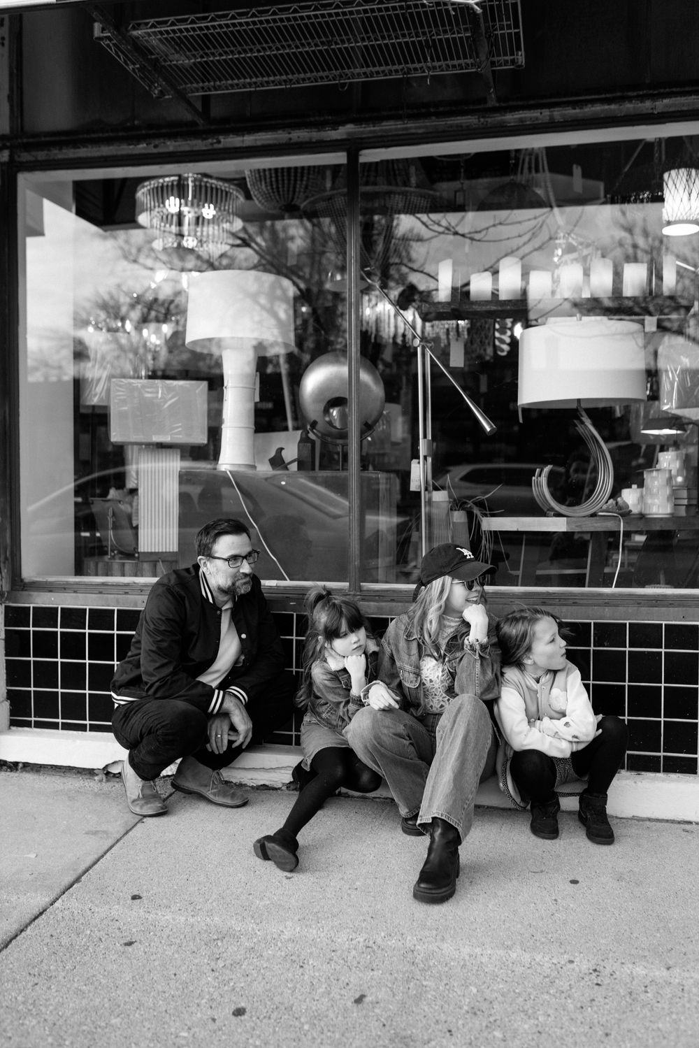 A group sits outside a storefront window with vintage lamps on display, captured in black and white photography.