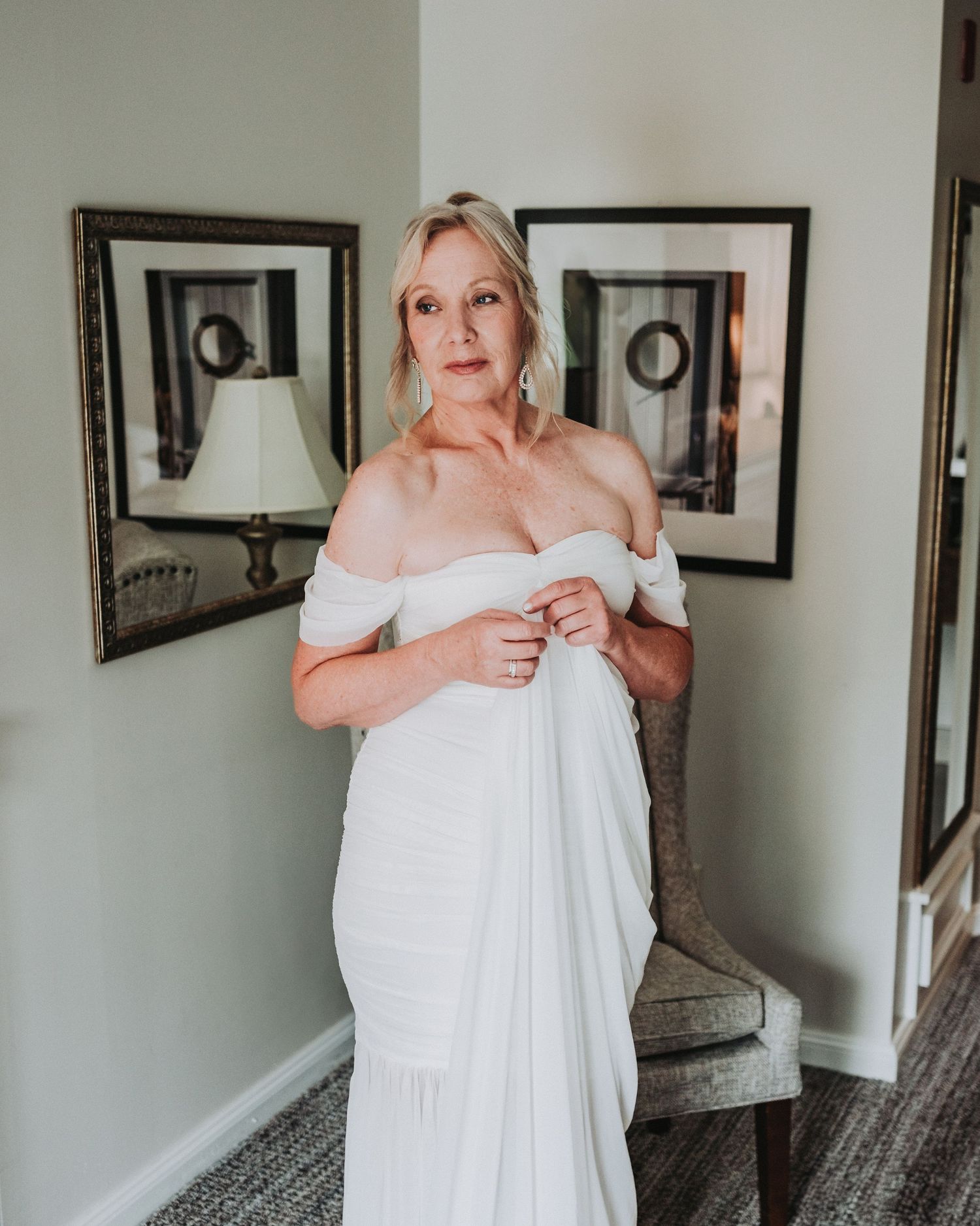 A bride in an elegant off-shoulder white wedding dress stands in front of framed artwork and a mirror in a room.
