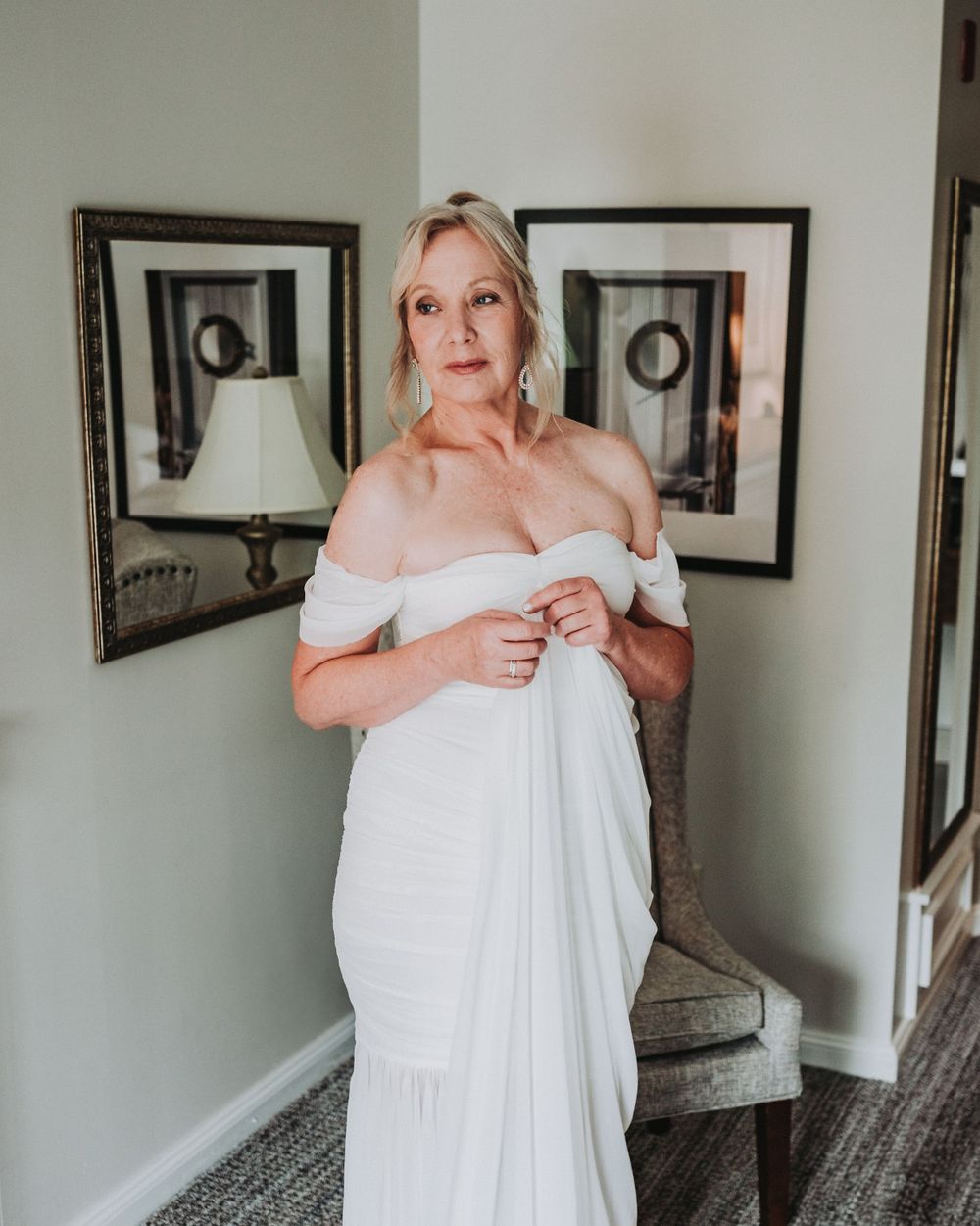 A bride in an elegant off-shoulder white wedding dress stands in front of framed artwork and a mirror in a room.
