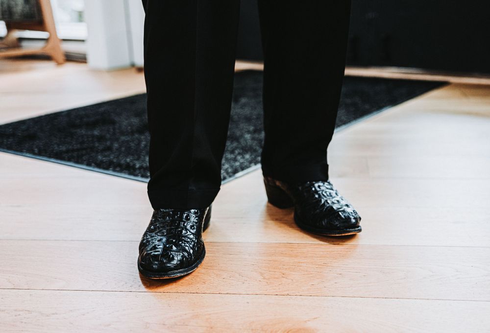The groom's black dress shoes with sparkly sequins stand out against light wooden flooring and a dark rug.