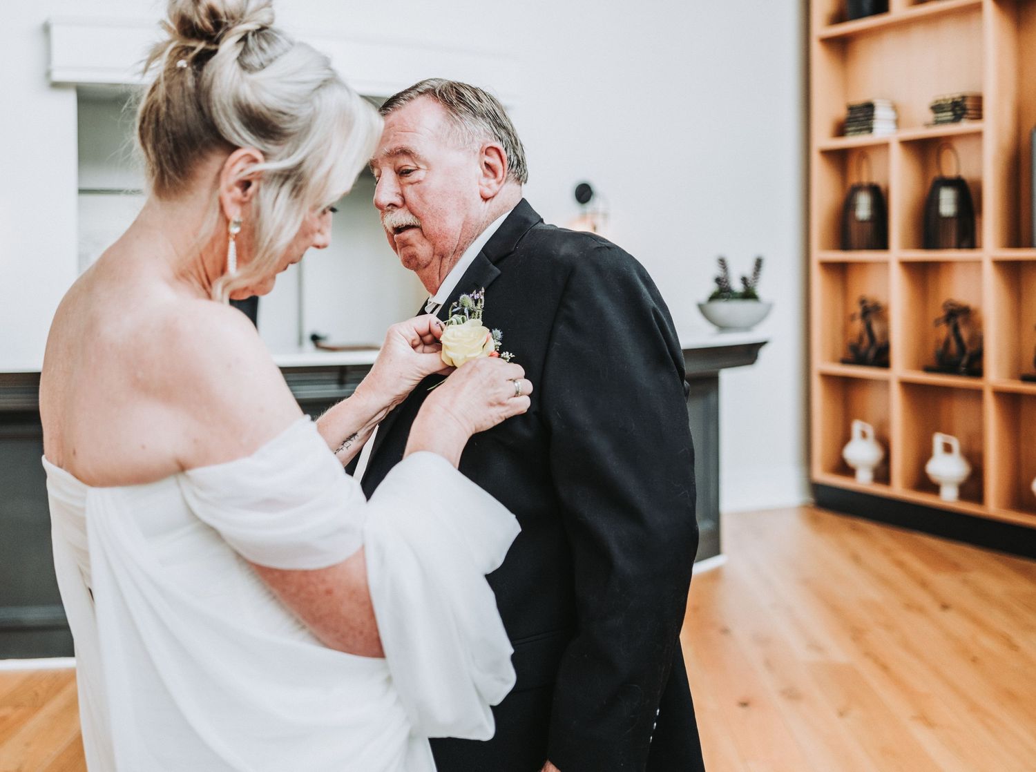 A tender moment during a wedding ceremony as a boutonniere is pinned on a black tuxedo jacket in a warm wooden interior.