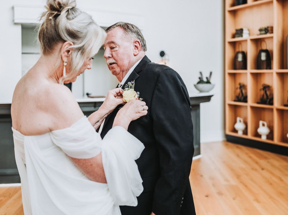 A tender moment during a wedding ceremony as a boutonniere is pinned on a black tuxedo jacket in a warm wooden interior.