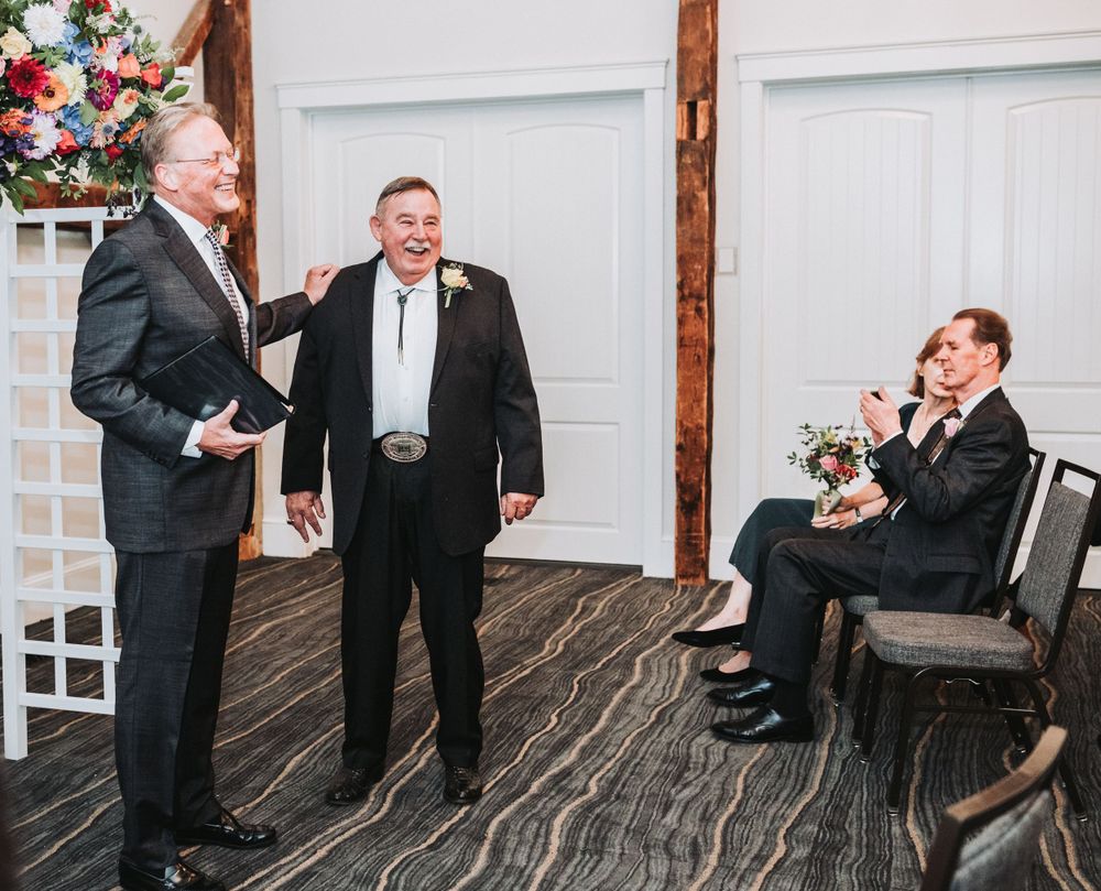 Three people share a lighthearted moment together in a rustic venue with wooden floors and floral decorations.