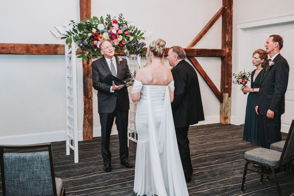 Wedding ceremony takes place under a floral arch with white and pink flowers in a rustic indoor venue.