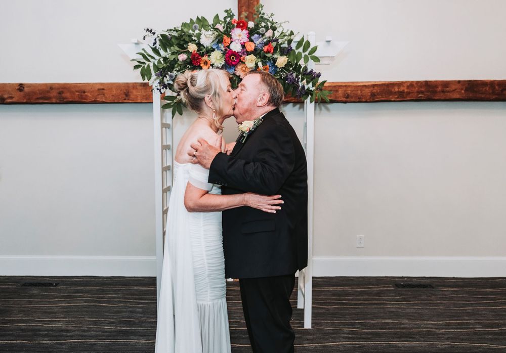 A couple embraces under a floral wedding arch with red and white flowers against a white wall and wooden floor.