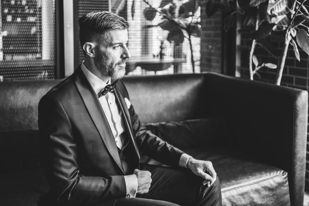 Black and white photo of a well-dressed person in a suit sitting pensively on a leather couch by a window.
