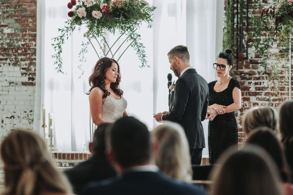 A couple exchanges vows during an indoor ceremony in front of a floral arch and exposed brick wall.