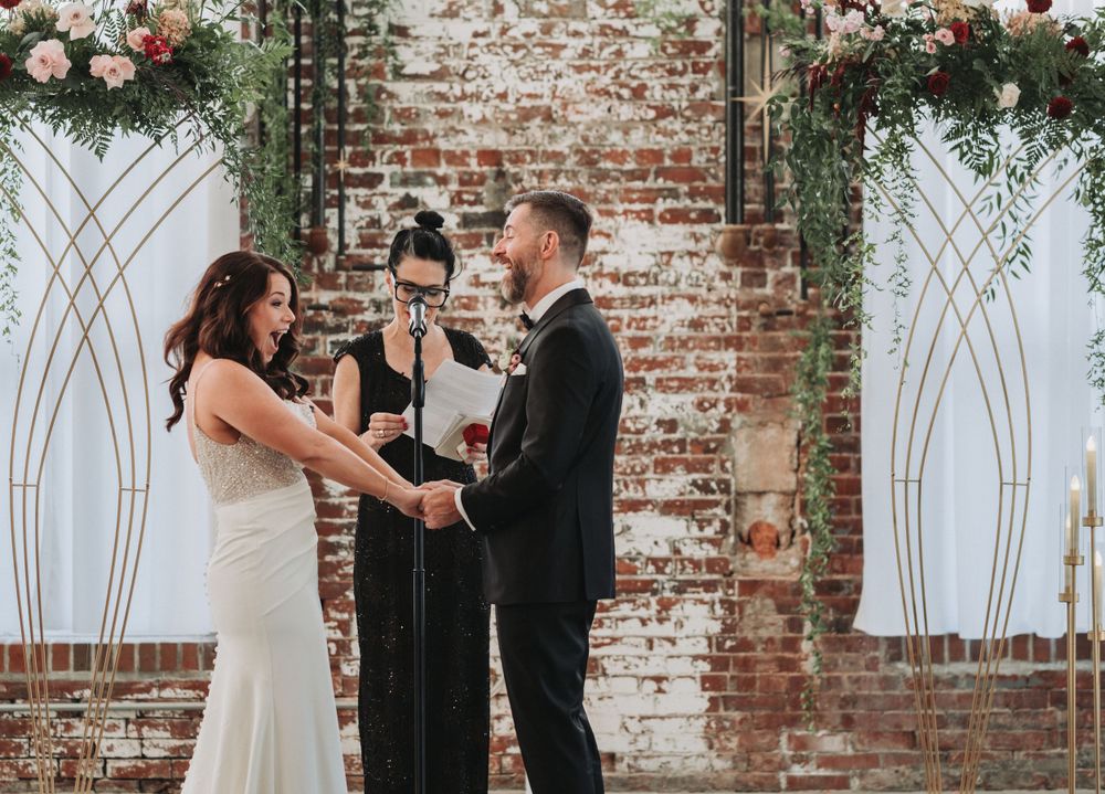 A wedding ceremony takes place in front of a rustic brick wall decorated with floral arrangements and geometric white arches.