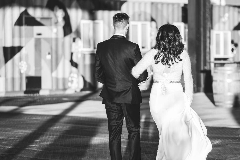 Black and white photo of a couple walking away from camera in formal wedding attire through a sunlit corridor.