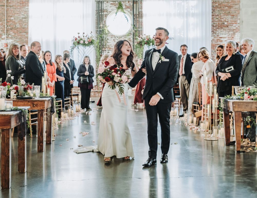 A joyful wedding couple walks down the aisle in a rustic industrial venue with exposed brick walls and gathered guests.
