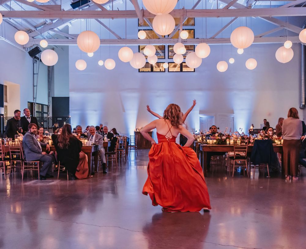 A maid of honor in a flowing red dress twirls at a reception venue decorated with glowing paper lanterns and elegant table settings.