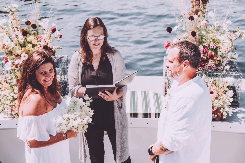 An outdoor wedding ceremony takes place with an officiant in black and a couple surrounded by floral arrangements.