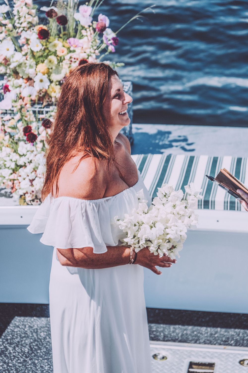 A bride in white off-shoulder dress holding white flowers on a boat deck with floral decorations and blue water background.