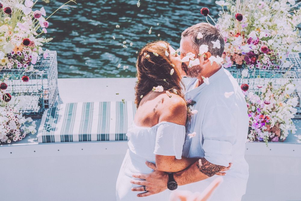 A romantic wedding moment captured on a boat deck with pink and purple floral decorations against sparkling water.