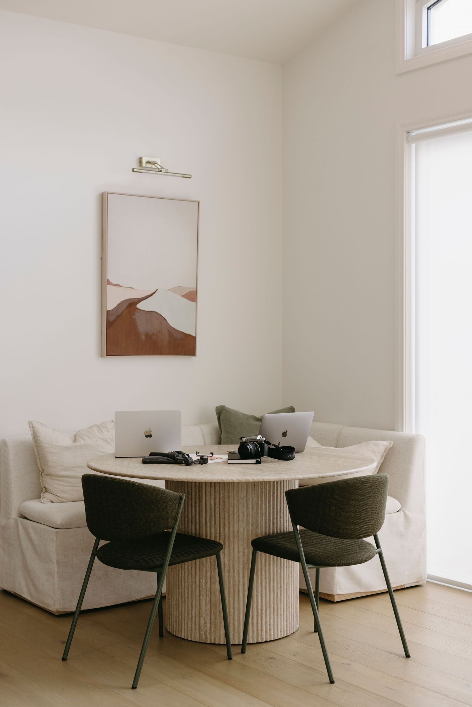 Modern minimalist dining nook with round fluted table, dark green chairs, cream sofa, and abstract wall art.