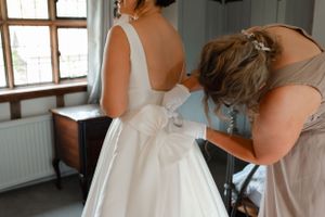 Someone helps adjust a bow detail on an elegant white wedding dress during bridal preparations in a sunlit room.