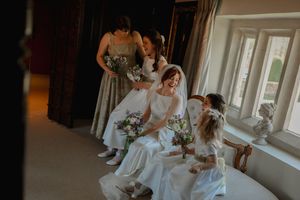 Wedding party in sage green bridesmaid dresses sitting by bright windows with bouquets at elegant venue.