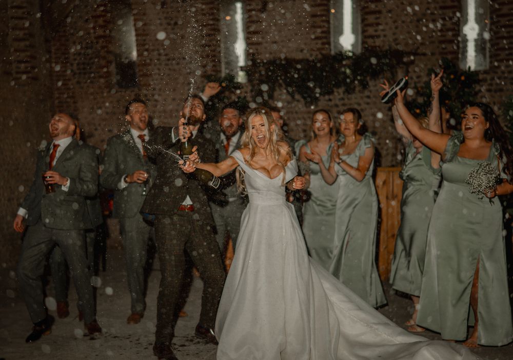 Wedding party in sage green dresses and gray suits celebrates joyfully as snow falls during evening reception.