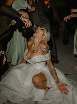 A person in a white wedding dress tilts back while someone pours champagne into their mouth at a dimly lit reception.