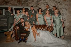 Wedding party poses together on vintage leather couch in rustic brick room with moody lighting and sage green attire.