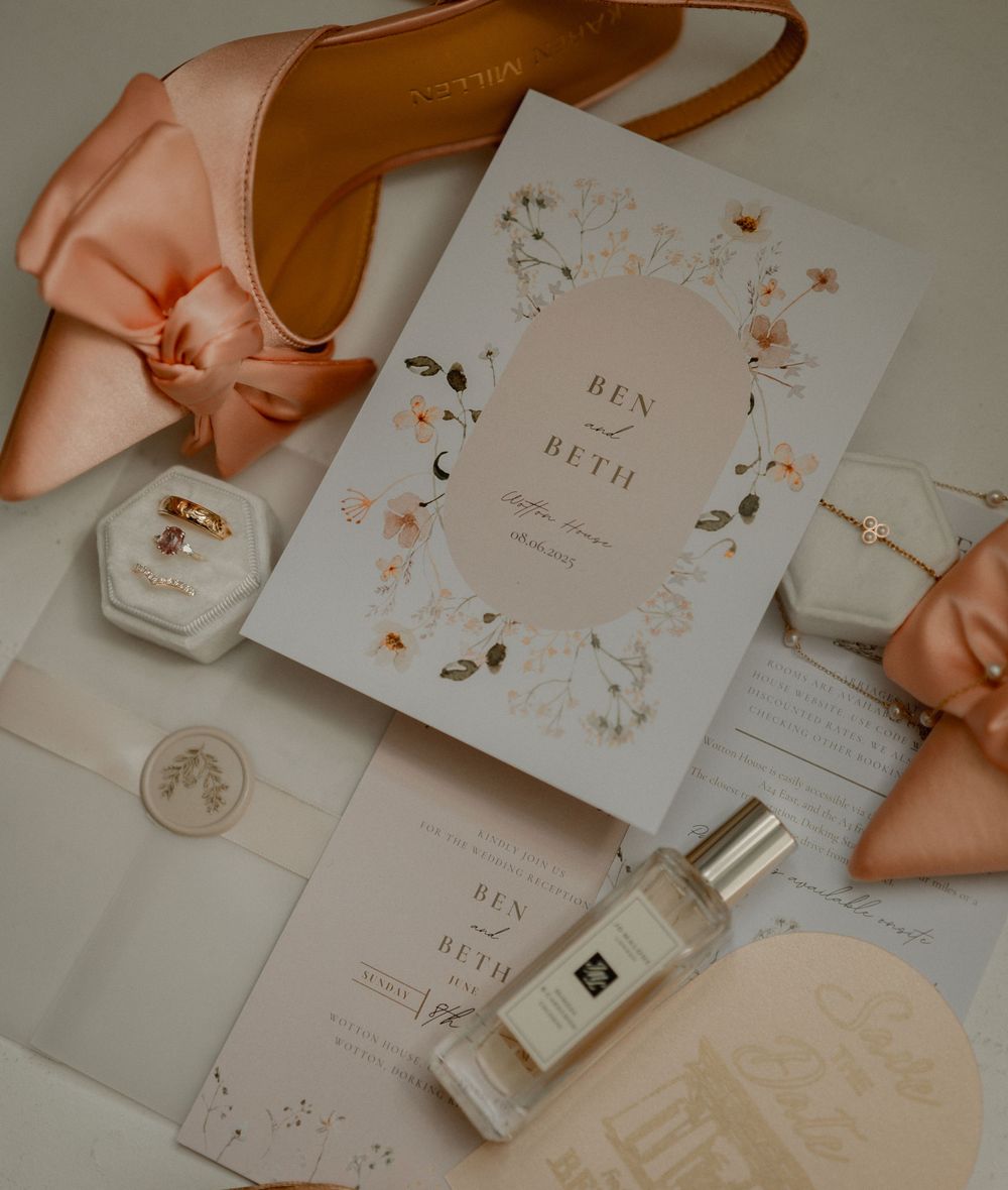 Wedding invitation with floral design in rose gold tones alongside perfume bottle, pink shoes and delicate accessories.