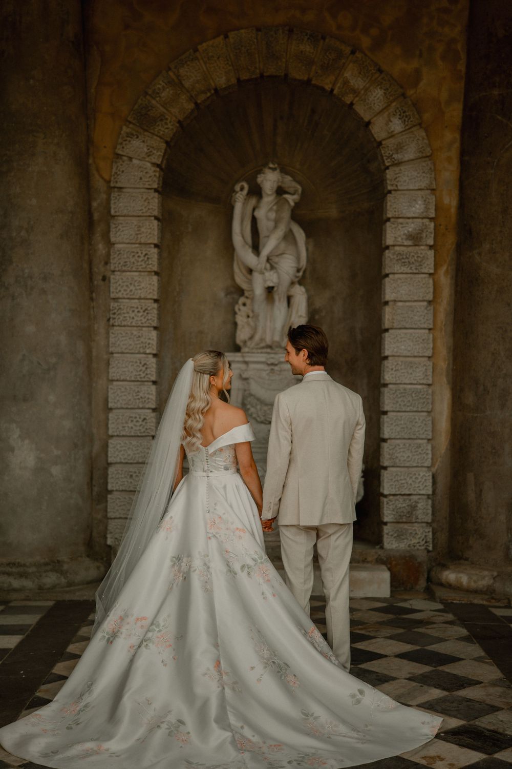 A wedding couple stands before a stone archway with a classical statue, the bride wearing an elegant off-shoulder gown with train.