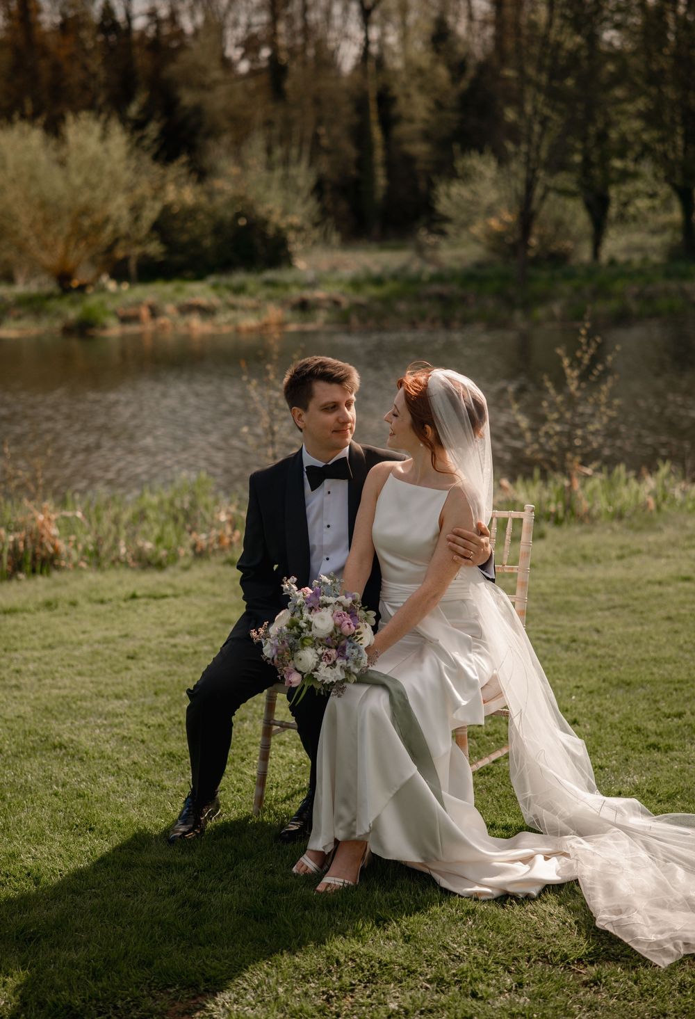 A wedding couple sits together on a rustic wooden bench by a serene lake surrounded by evergreen trees.