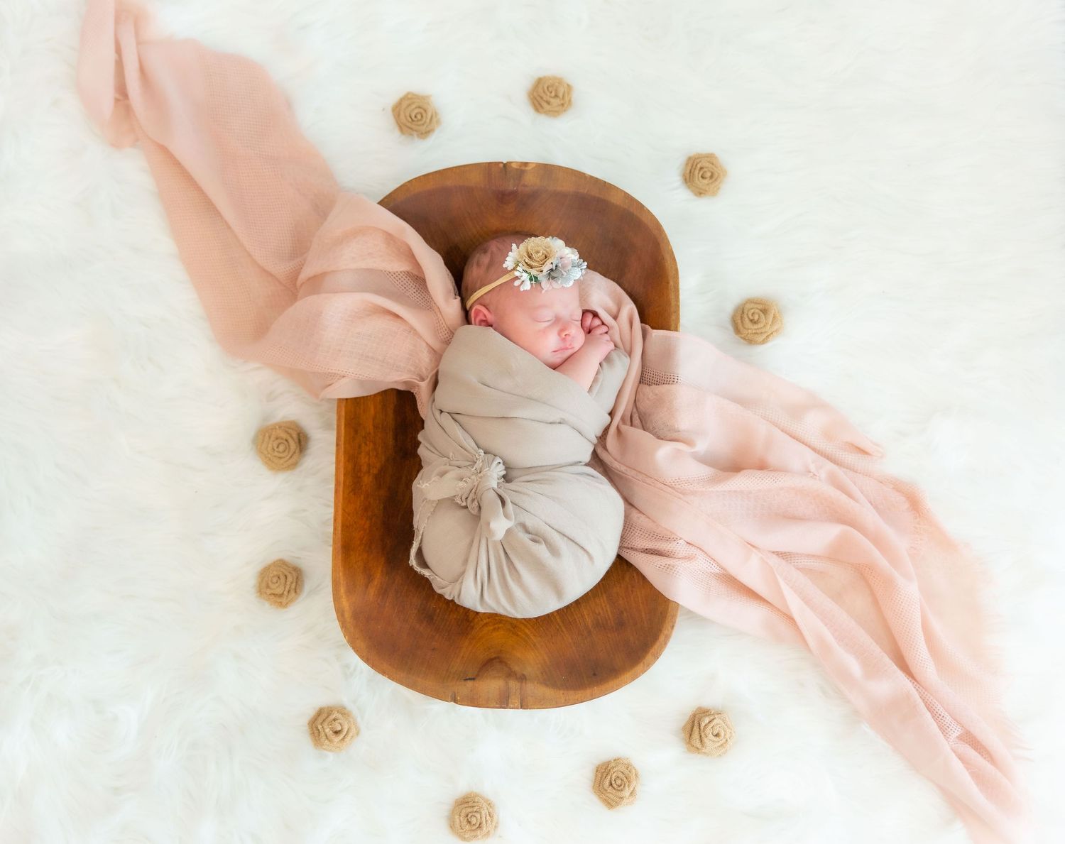Newborn photography with baby in wooden bowl surrounded by pink fabric and decorative elements.