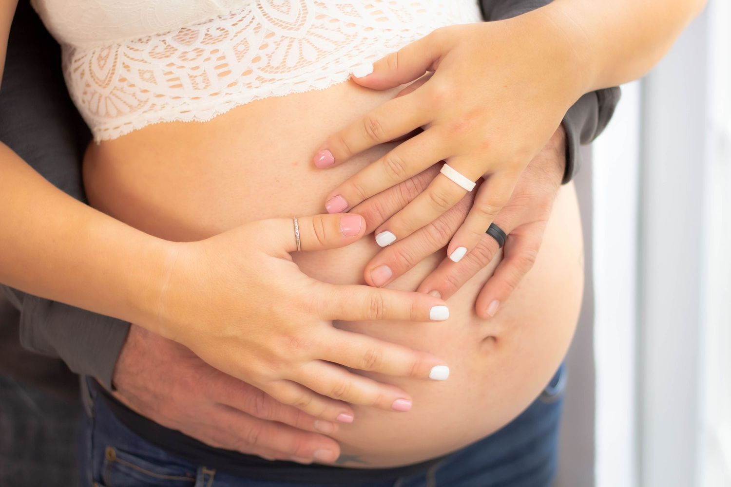 Hands lovingly rest on a pregnant belly wearing blue jeans and white lace.