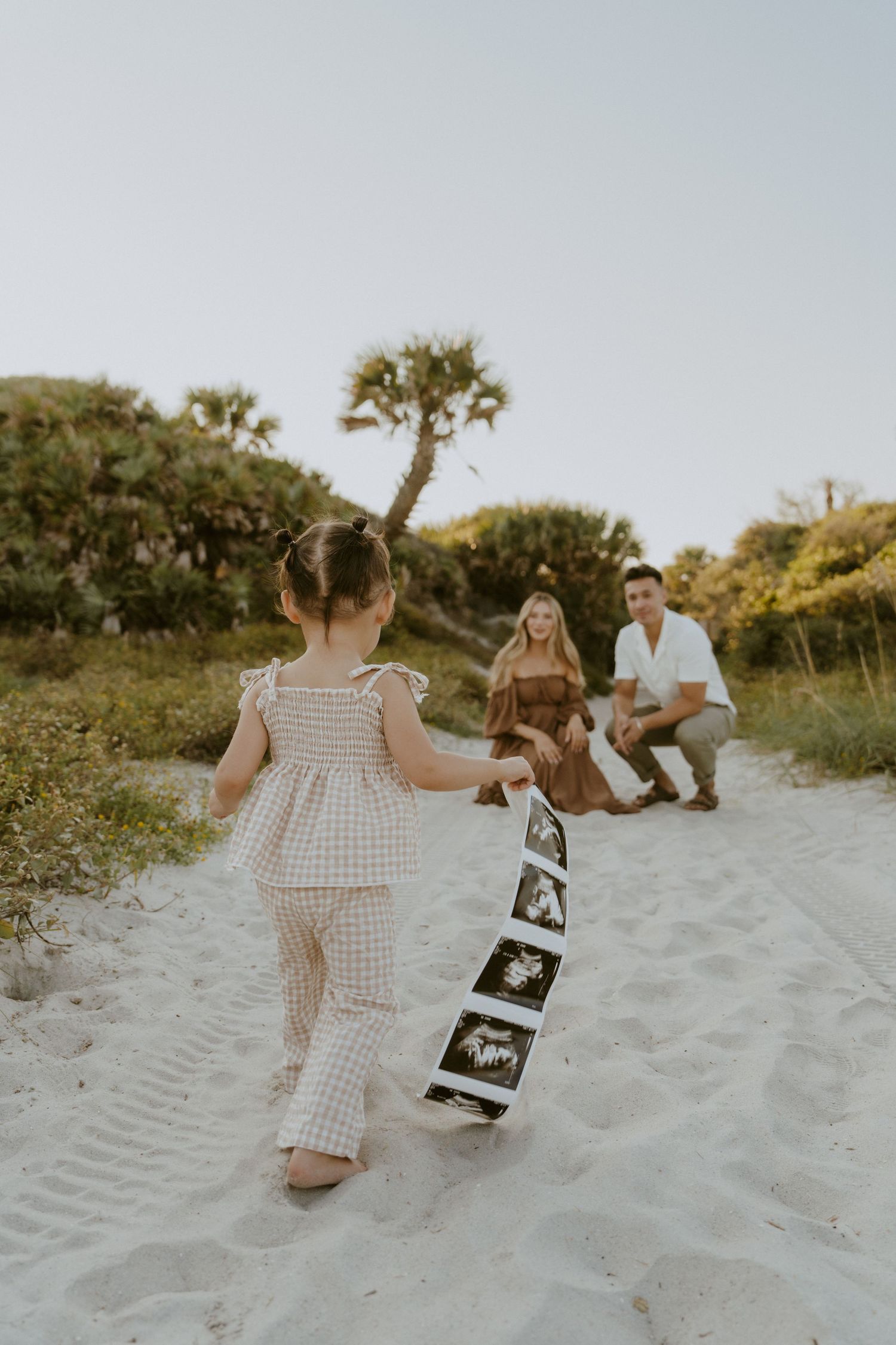 A child walks on beach sand holding ultrasound photos toward adults sitting in background.