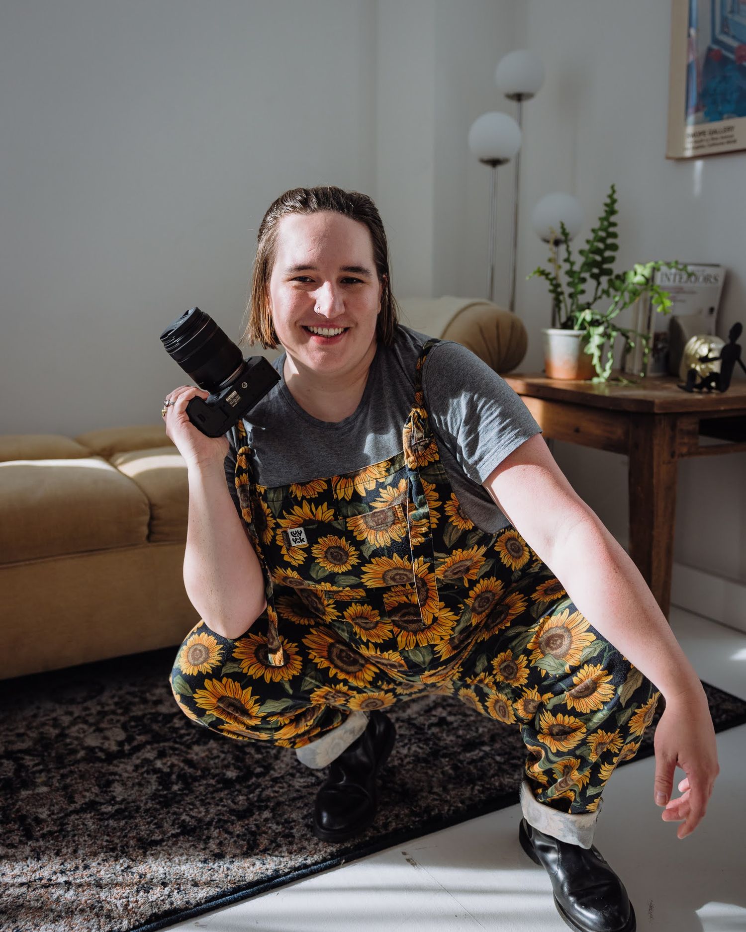 A smiling photographer in sunflower overalls poses with a camera in a cozy indoor setting.