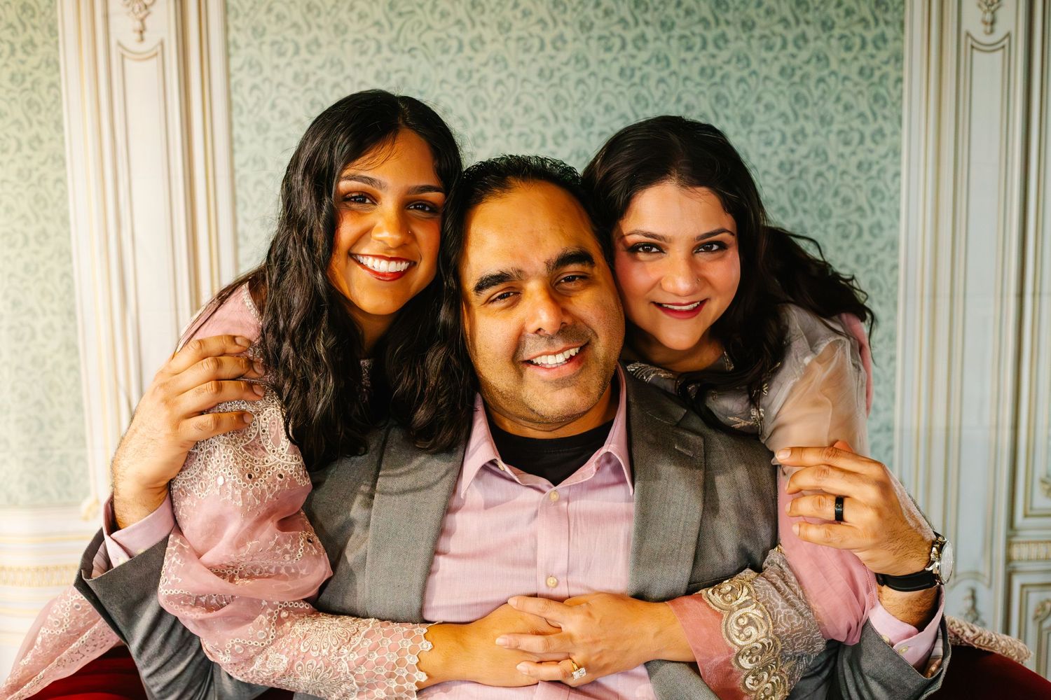 Family smiling warmly at the camera in studio while sitting together in a room with patterned wallpaper with Nichole Sieck Photography.