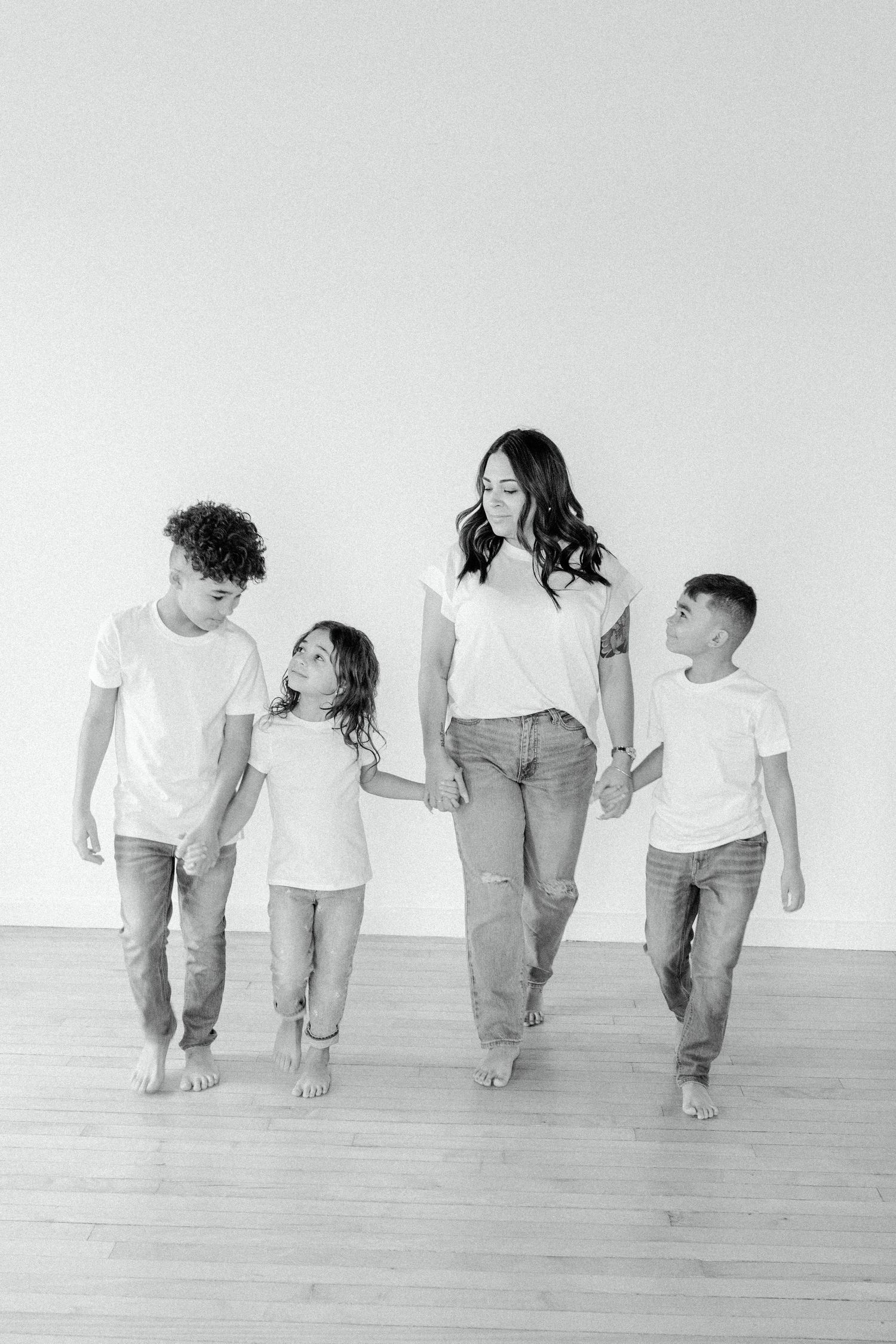 A family walks together barefoot in a minimalist white studio while holding hands and wearing casual outfits.