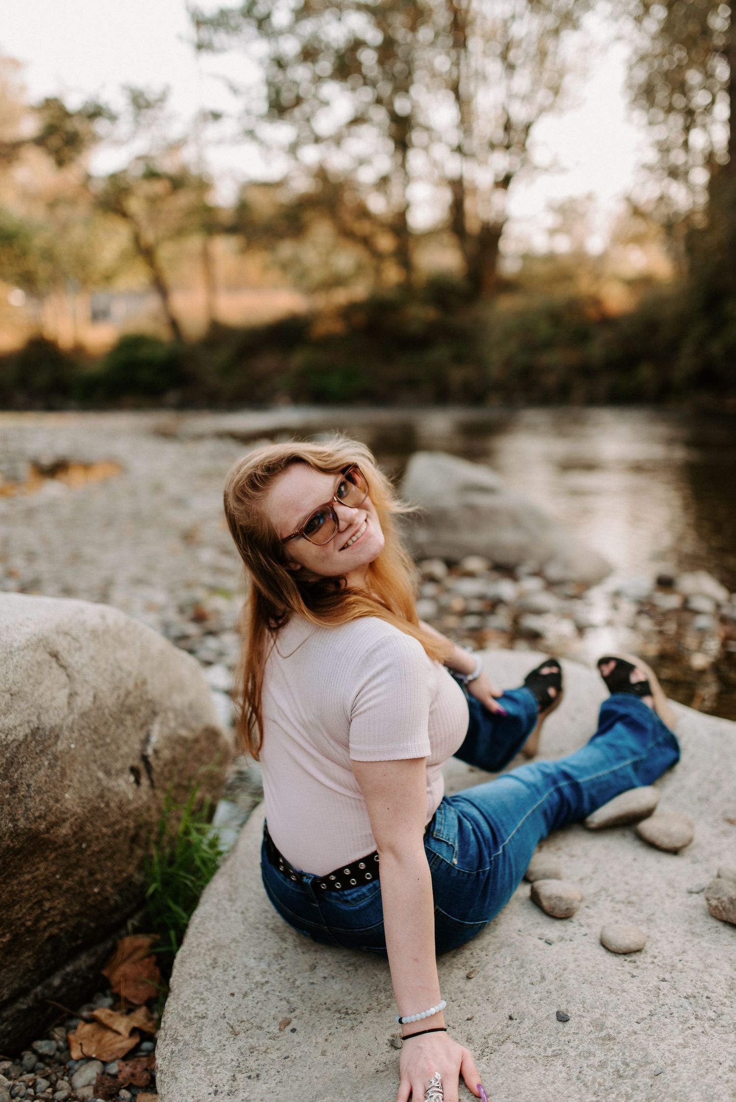 Person relaxing on large rock by river during golden hour wearing casual attire.