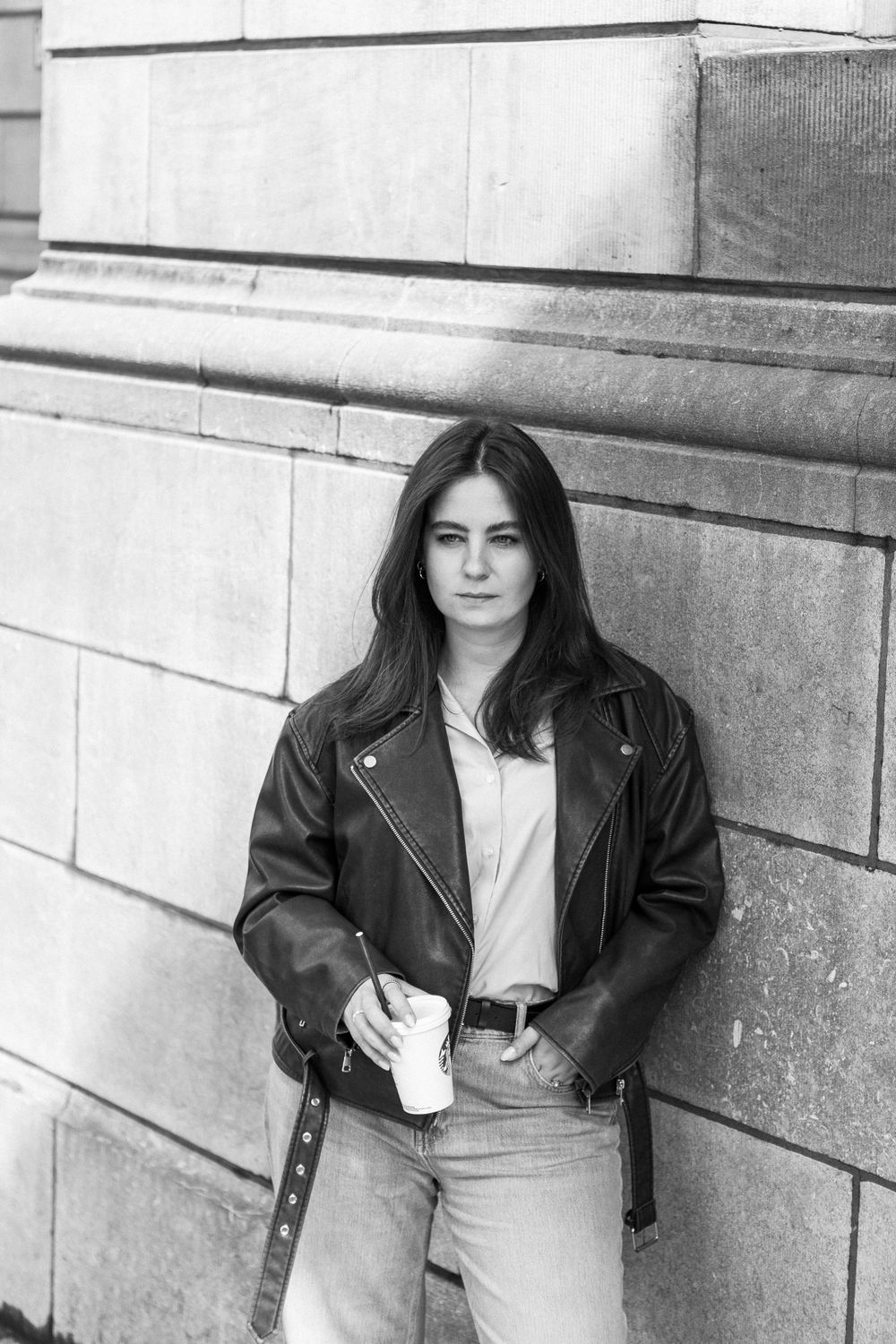 Black and white portrait of person in leather jacket leaning against stone wall.