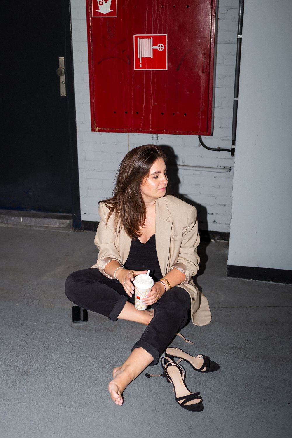 Fashion model in beige blazer and black pants sitting casually by red fire safety sign.