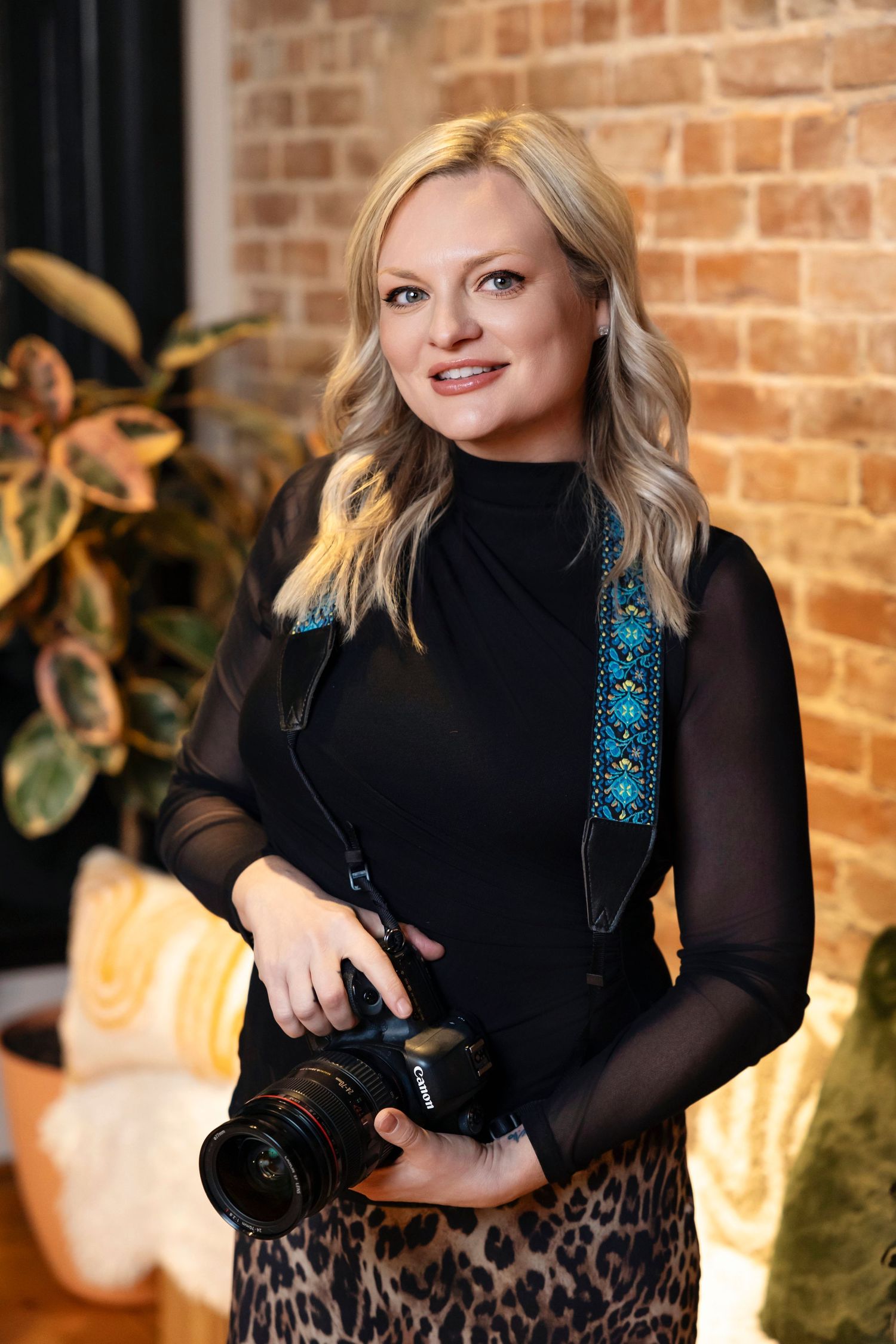 Professional photographer in black sheer top and leopard print skirt holding camera against brick wall background.