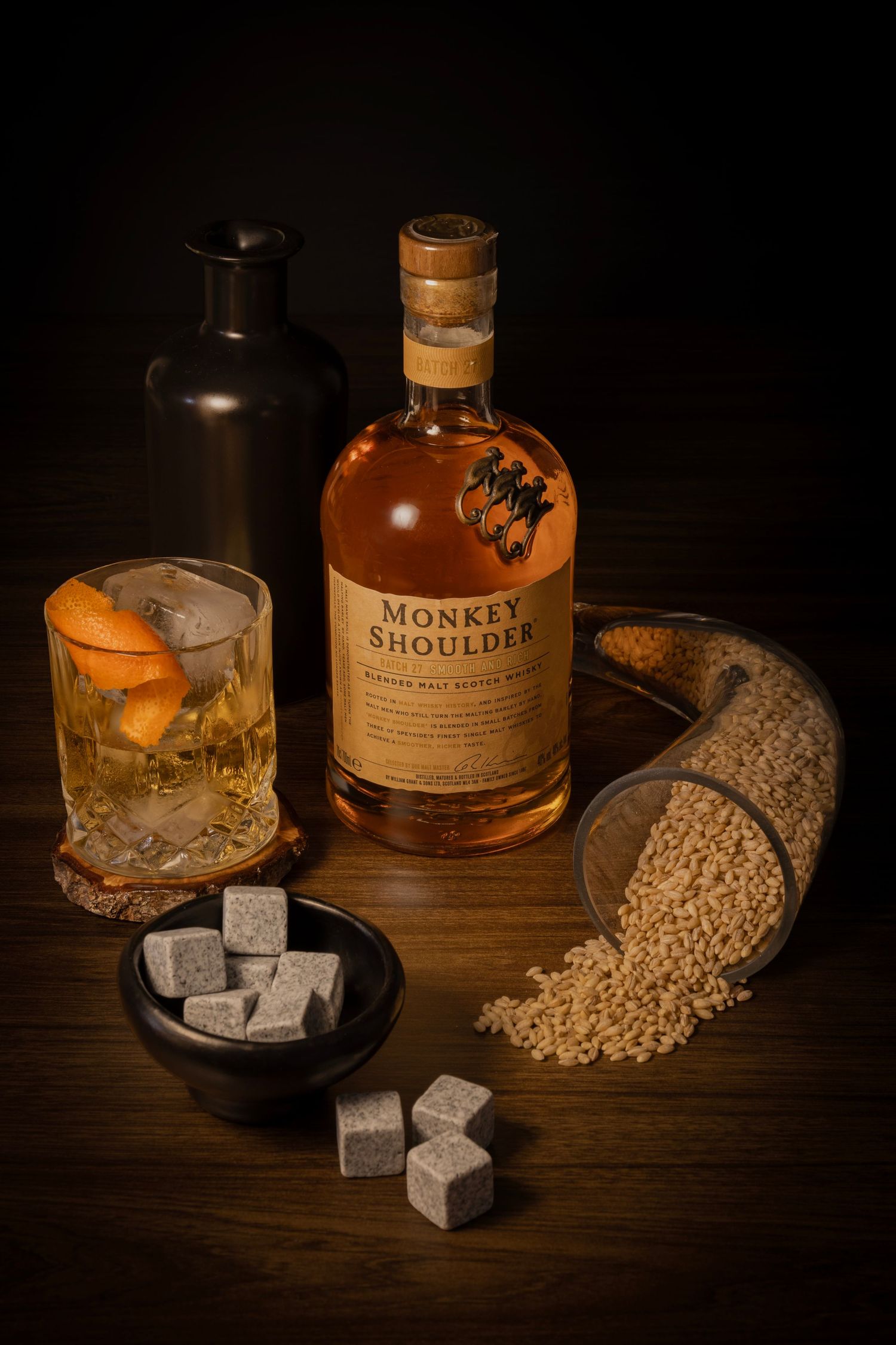 Whisky with orange peel, bottle, black jug, spilled barley, and whisky stones on a wooden surface in warm lighting.