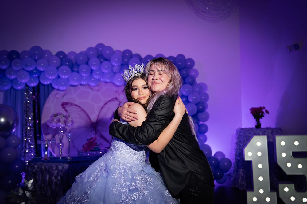 Two people share an emotional embrace at a quinceañera celebration with purple balloon decorations.