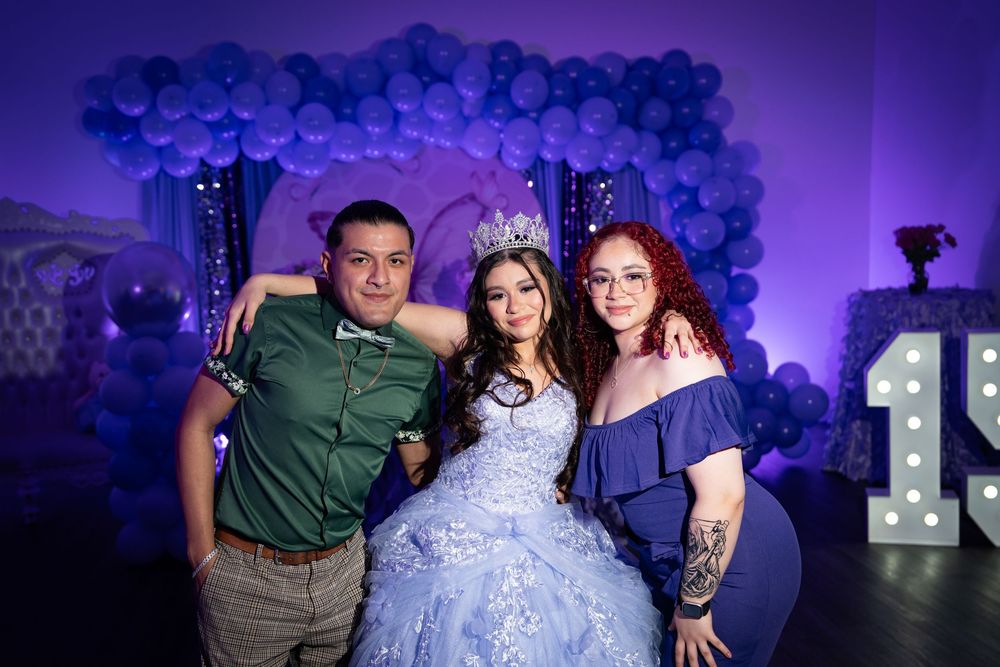 Three people pose together at a formal celebration with blue balloon decorations and light-up numbers.