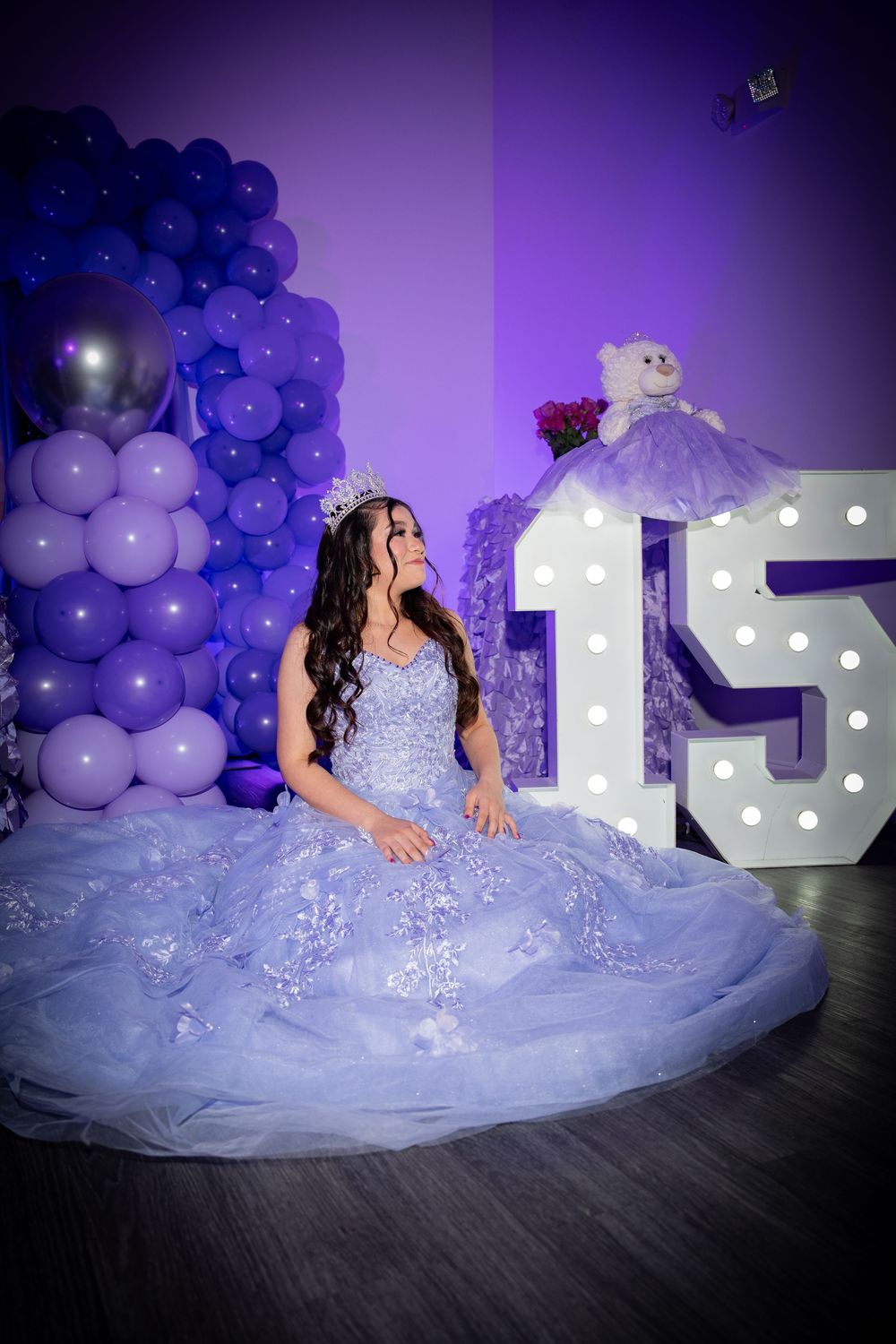 Person in a lavender ball gown poses by number 15 display with purple balloon arch backdrop.